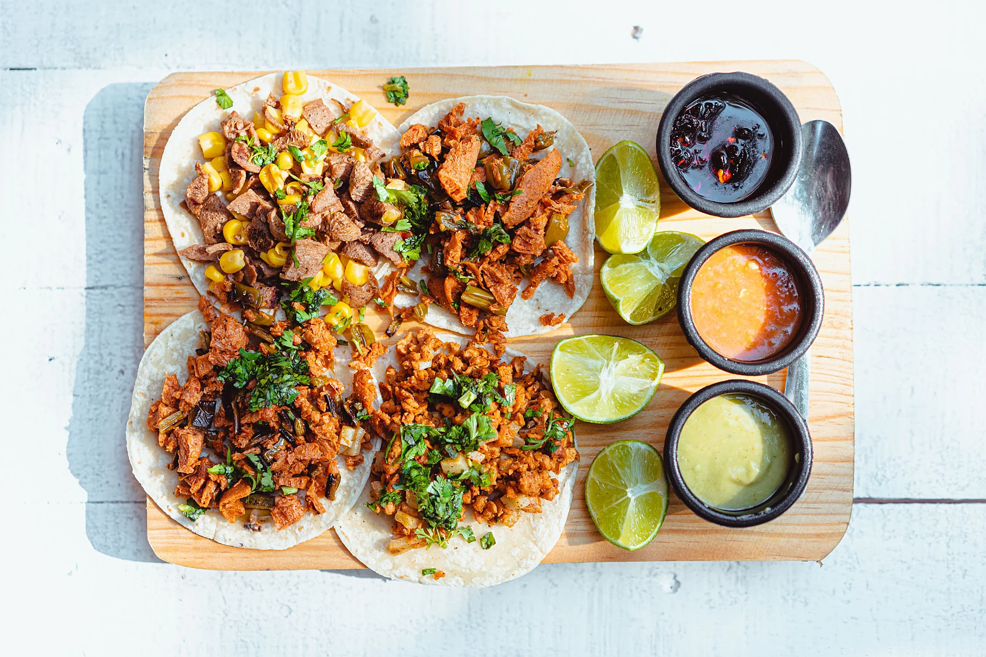 Four street tacos with lime and three salsas on a wooden board